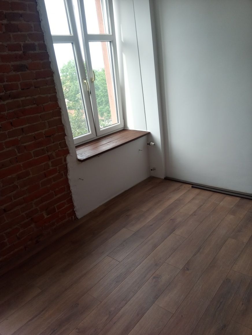 Wnętrze pokoju w trakcie prac wykończeniowych: ułożone panele podłogowe, fragment ściany z cegły, okno z drewnianym parapetem, biała ściana.
