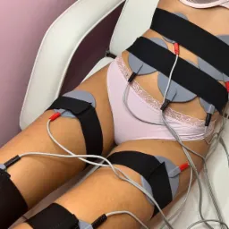 Kobieta w bieliźnie poddawana zabiegowi elektrostymulacji mięśni brzucha i ud z użyciem elektrod i pasów mocujących w gabinecie kosmetycznym.