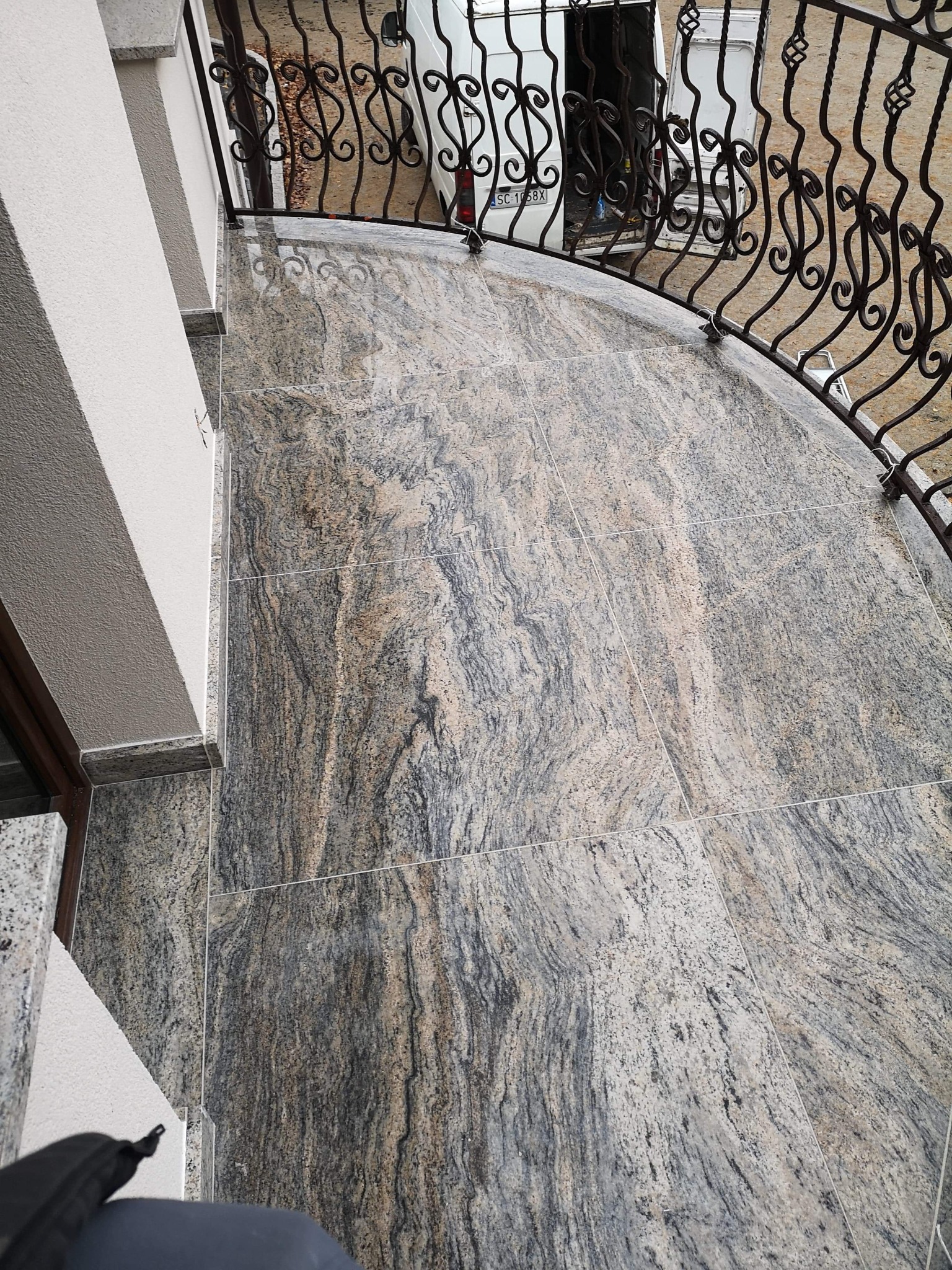 Wykończony balkon z kamiennymi płytami o wzorze przypominającym marmur, z ozdobną, kutą balustradą i widocznym fragmentem białej ściany budynku oraz zaparkowanego w tle białego samochodu dostawczego.