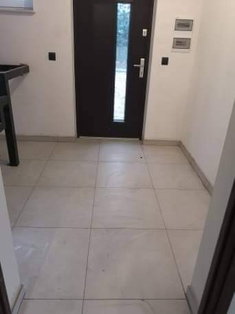 Wyłożona jasnymi, kwadratowymi płytkami podłoga w przedpokoju z ciemnymi drzwiami wejściowymi z przeszkleniem; widoczne detale wykończenia przy ścianach i fragment mebla po lewej stronie.