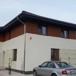 Dwukondygnacyjny dom z brązową drewnianą elewacją na górze i kremową na dole, widoczny numer domu '4' i srebrny samochód zaparkowany przed budynkiem.