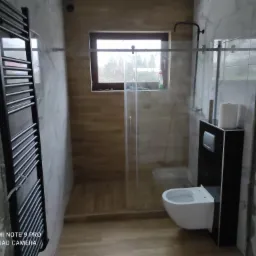 Łazienka w trakcie remontu: widoczne płytki imitujące drewno w kabinie prysznicowej, częściowo wykończone marmurowe ściany, czarny grzejnik drabinkowy, wisząca toaleta z czarną zabudową i okno...