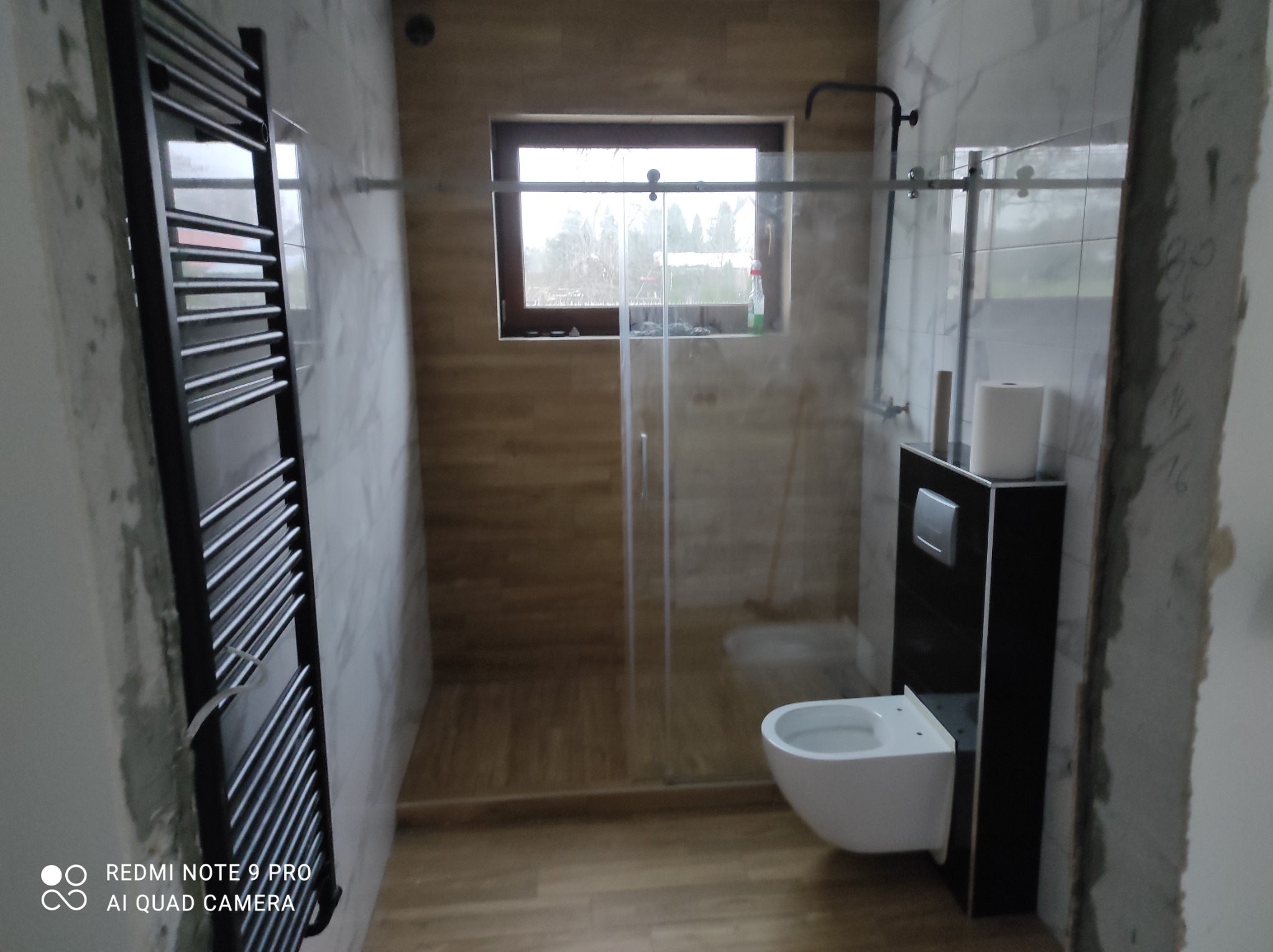 Łazienka w trakcie remontu: widoczne płytki imitujące drewno w kabinie prysznicowej, częściowo wykończone marmurowe ściany, czarny grzejnik drabinkowy, wisząca toaleta z czarną zabudową i okno...