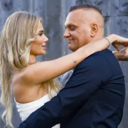 Para młoda obejmująca się na tle szarej drewnianej ściany; panna młoda w białej sukni z odkrytymi ramionami i tatuażem na ramieniu, pan młody w ciemnym garniturze.