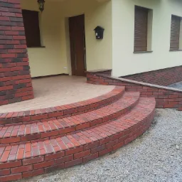 Wykończone schody wejściowe do domu z czerwonej cegły klinkierowej, z kolumną z tego samego materiału i żwirowym podłożem.