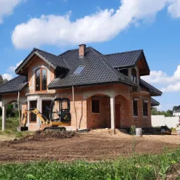 Budowa domu w stanie surowym otwartym, widoczna mini koparka na działce przed budynkiem, przygotowany teren pod dalsze prace.