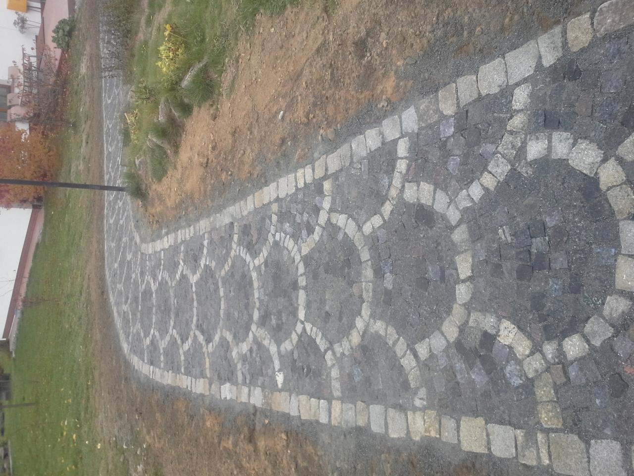 Ścieżka z kostki granitowej ułożona we wzór z jasnymi obwódkami na trawiastym terenie.