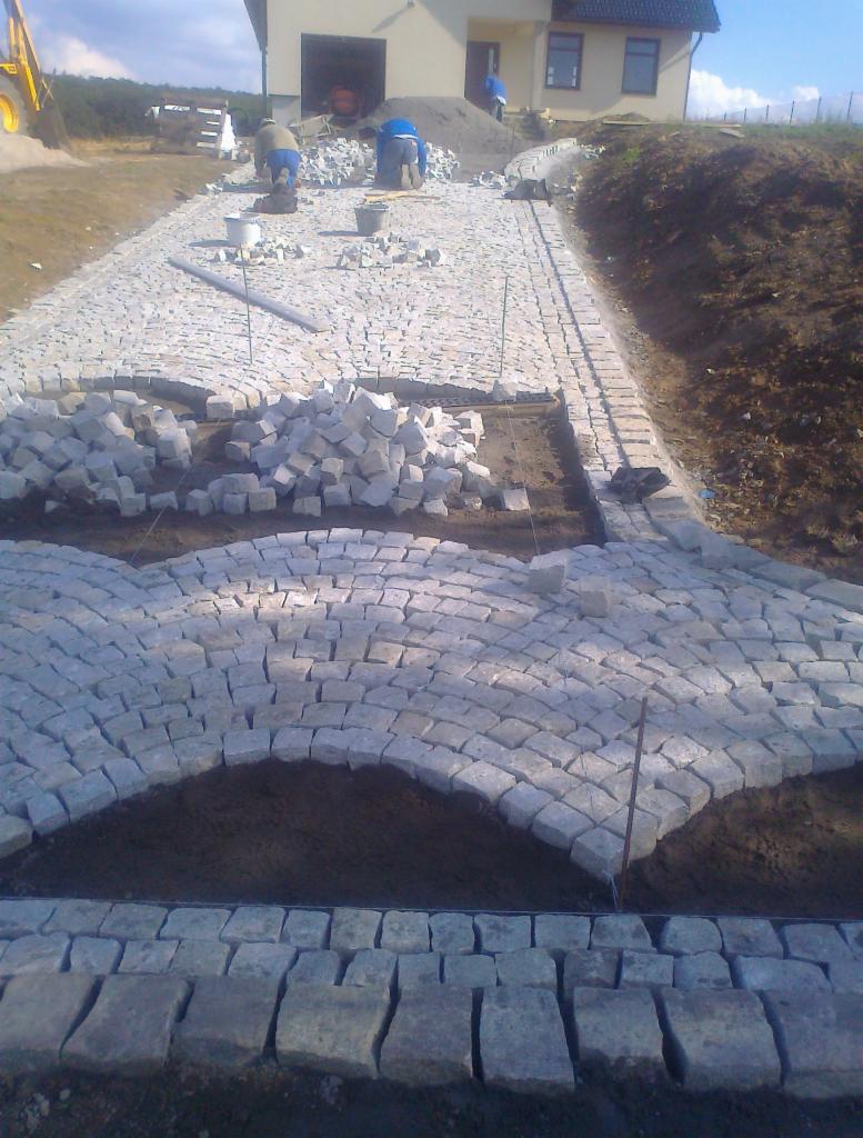 Trwa układanie wzoru z szarej kostki na podjeździe, widoczni robotnicy i sterta kostki do ułożenia, dom w tle.