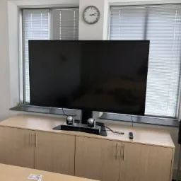 Duży telewizor z wideokonferencyjnym zestawem głośnomówiącym i kamerami na szafce w biurze, obok okna z żaluzjami i rośliną doniczkową, z widocznym zegarem na ścianie.