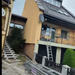 Prace na wysokości przy elewacji domu: mężczyzna na drabinie przy balkonie, widoczne rusztowania i narzędzia, budynek w trakcie prac wykończeniowych.