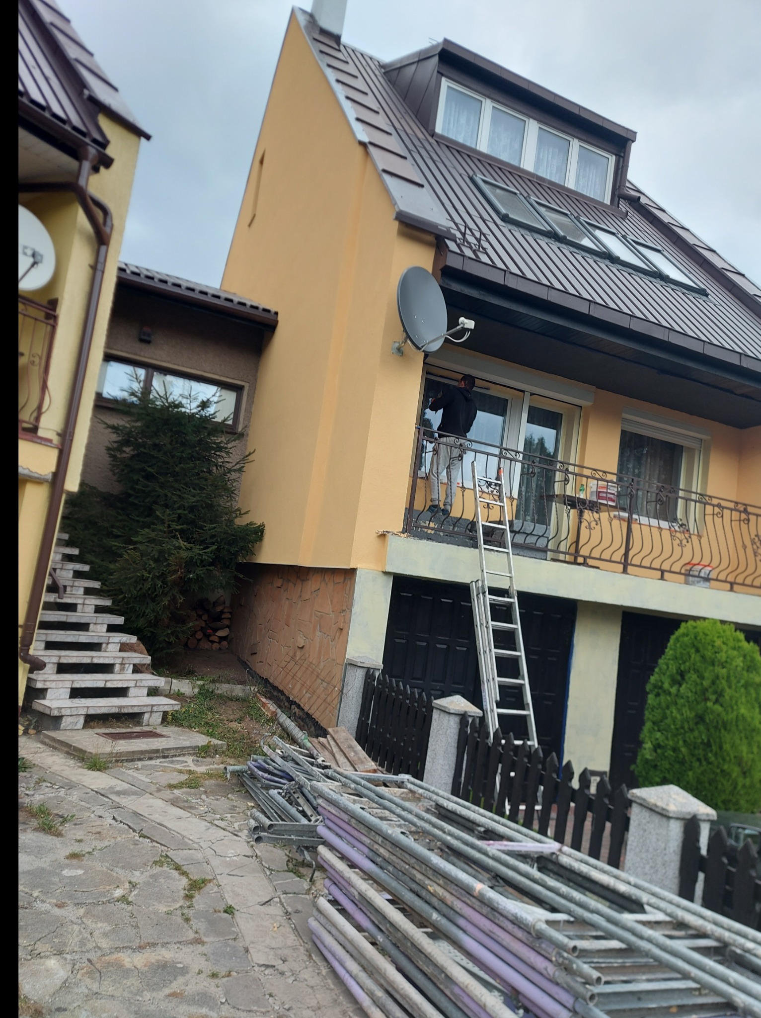 Prace na wysokości przy elewacji domu: mężczyzna na drabinie przy balkonie, widoczne rusztowania i narzędzia, budynek w trakcie prac wykończeniowych.