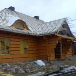 Dom z bali w trakcie budowy dachu, widoczne krokwie i membrana dachowa, okna zabezpieczone folią, teren wokół domu częściowo zagospodarowany kamieniami.