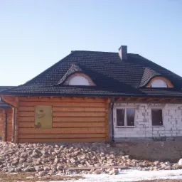 Dom w trakcie budowy: część murowana z oknami, część z bali, dach pokryty ciemną dachówką z lukarnami w kształcie półksiężyca, teren wokół usypany kamieniami.