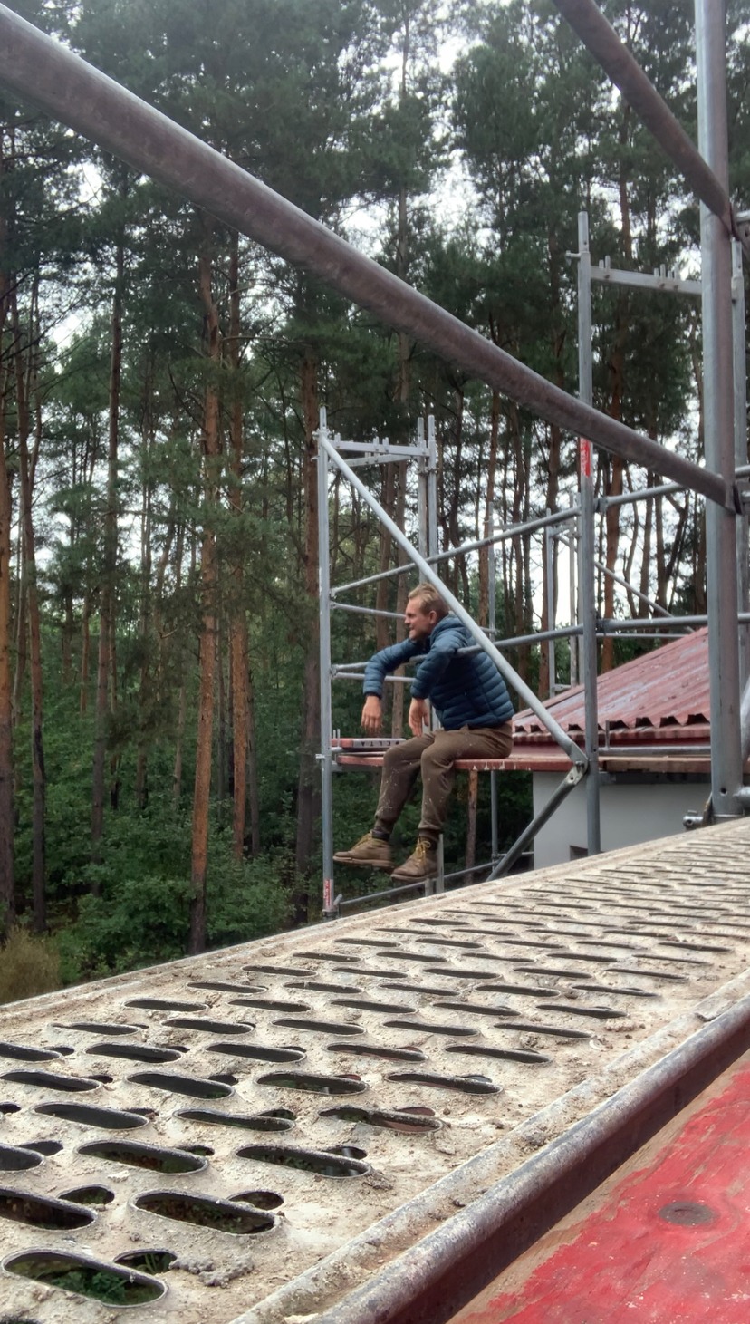 Pracownik w niebieskiej kurtce siedzi na rusztowaniu na tle lasu, z widocznym fragmentem dachu budynku.