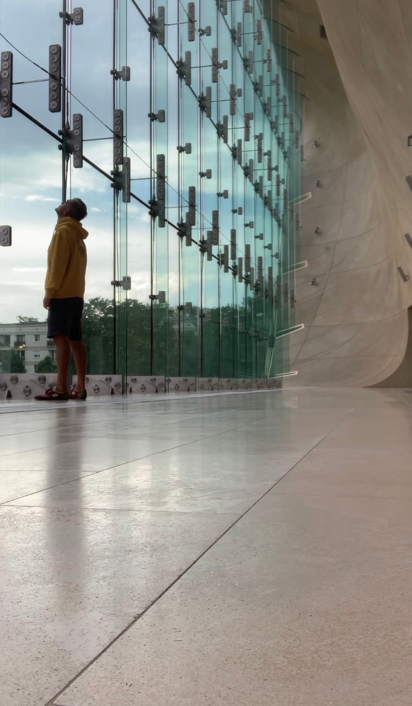 Wnętrze budynku z przeszkloną ścianą, widoczny mężczyzna w żółtej bluzie spoglądający w górę na konstrukcję mocowania szyb.