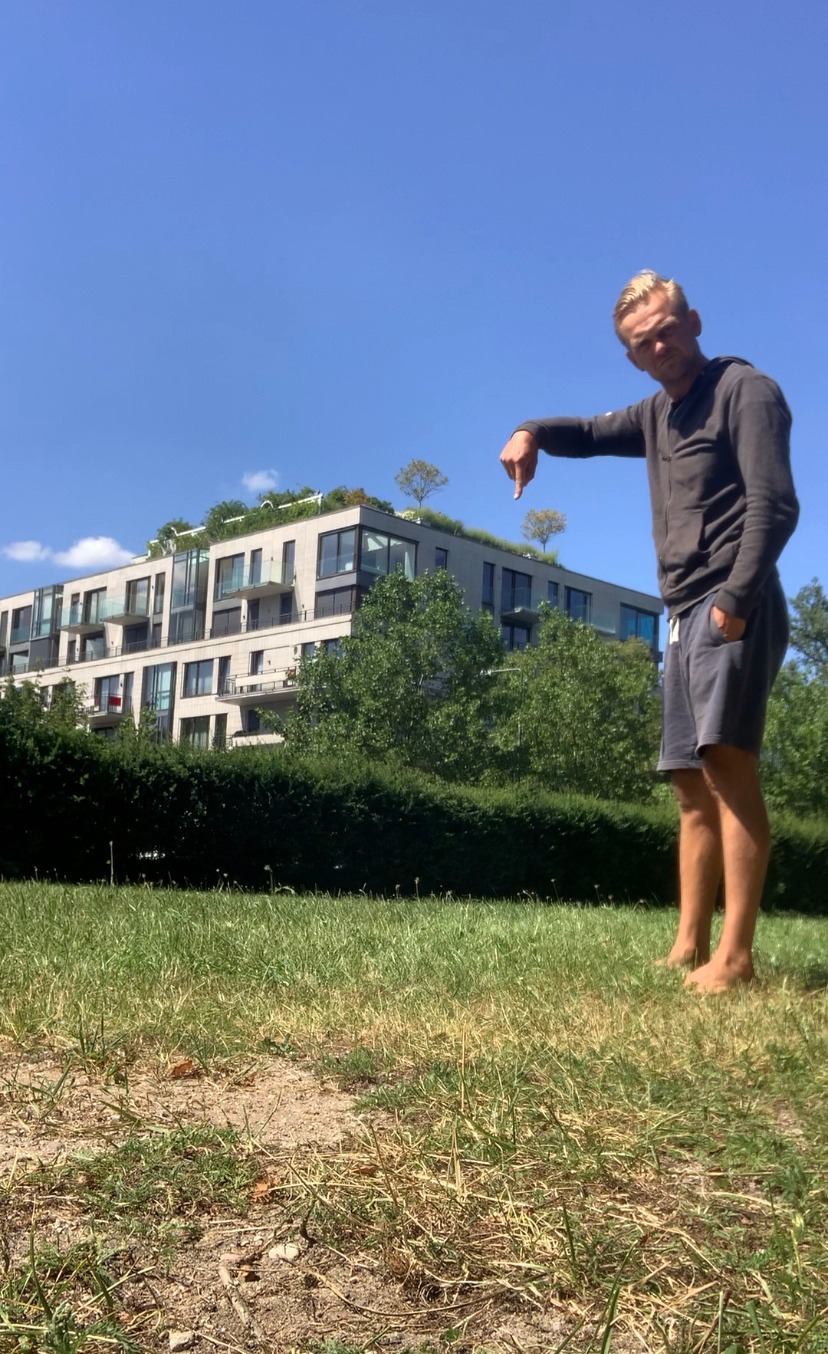 Mężczyzna bosymi stopami na trawie wskazuje palcem w dół, w tle nowoczesny budynek z zielonym dachem i drzewami.