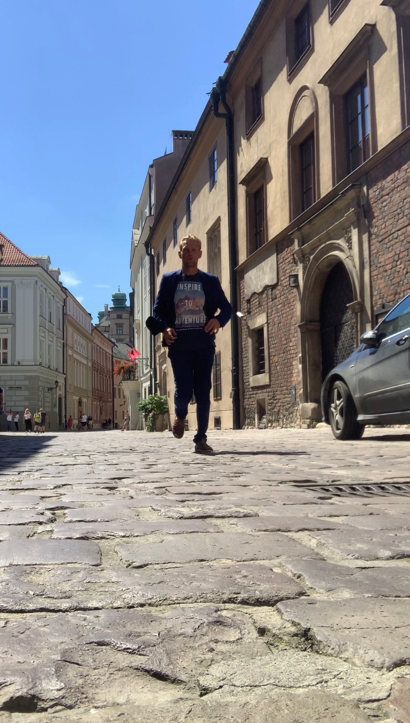 Mężczyzna w garniturze biegnie po brukowanej ulicy w historycznej części miasta, w tle kamienice z cegły i piaskowca, widoczne detale architektoniczne i błękitne niebo.