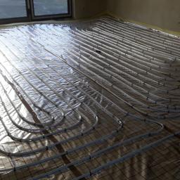 ESklima - Rozłożona instalacja ogrzewania podłogowego z rurkami na folii z siatką, widoczne okno i fragment ściany w surowym stanie.