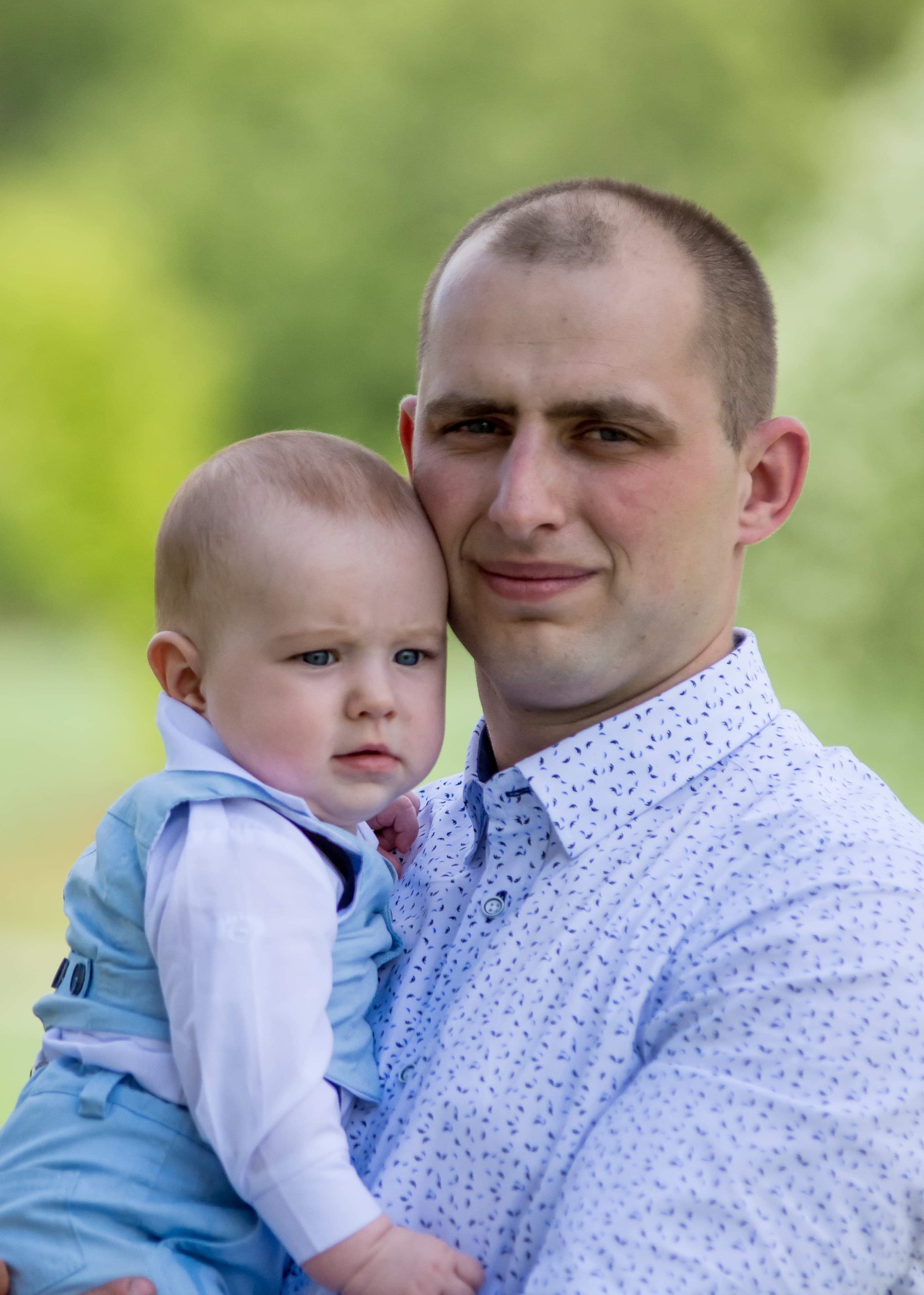 Mężczyzna w jasnej koszuli w drobny wzór trzyma na rękach małego chłopca ubranego w błękitny garnitur, oboje patrzą w obiektyw na rozmytym zielonym tle.