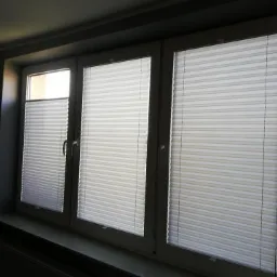 Trzyczęściowe okno z zamontowanymi białymi roletami plisowanymi, częściowo zasłoniętymi, na tle ciemnej ściany.