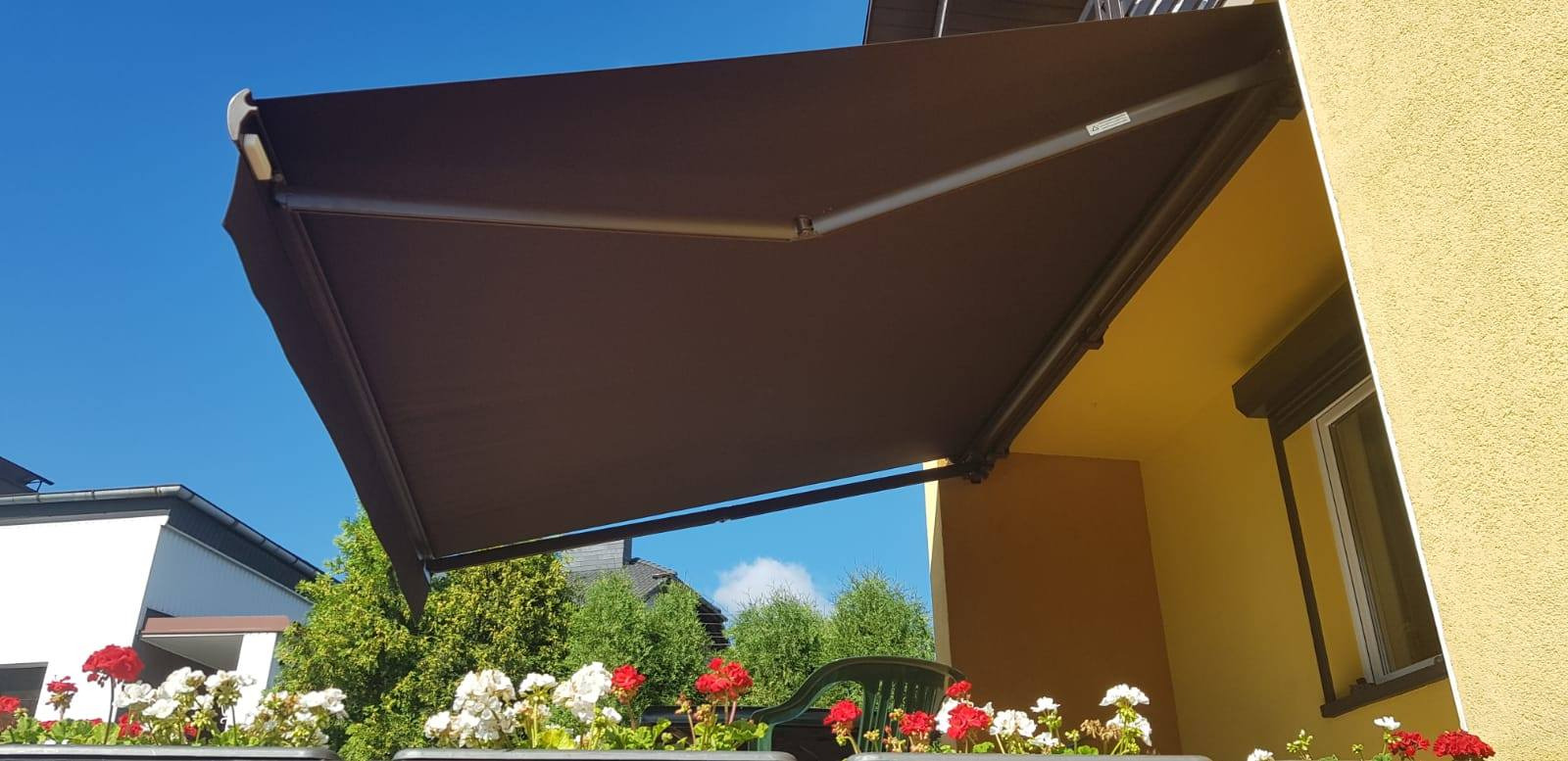 Rozwinięta, brązowa osłona przeciwsłoneczna nad balkonem, widok z dołu na tle błękitnego nieba i kwitnących kwiatów w donicach.