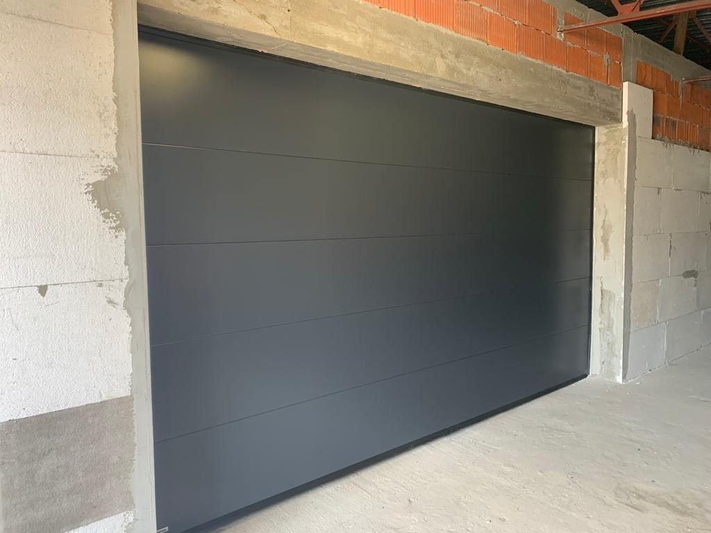 Zamknięta, segmentowa brama garażowa w kolorze antracytowym, widoczna na tle surowych ścian z betonu komórkowego i cegły.
