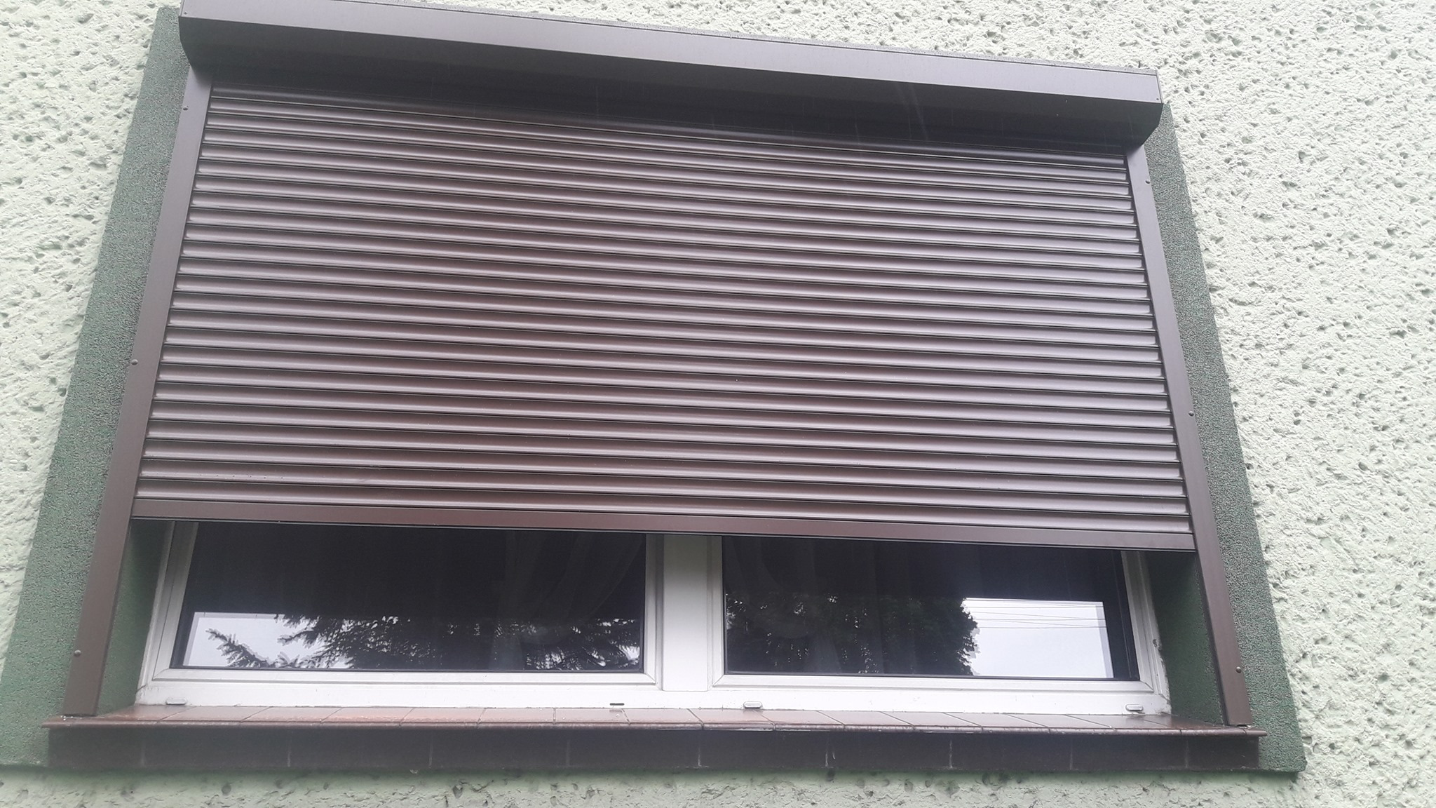 Ciemnobrązowa osłona okienna z częściowo opuszczonym pancerzem na tle zielonej, chropowatej ściany zewnętrznej budynku. Widoczne odbicie drzew w szybie okiennej.
