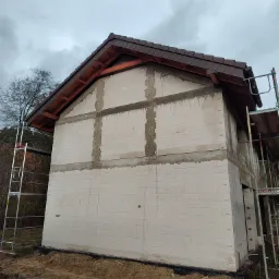 Dwukondygnacyjny dom w stanie surowym otwartym, z brązową dachówką i widoczną więźbą dachową, otoczony rusztowaniami i ziemią.