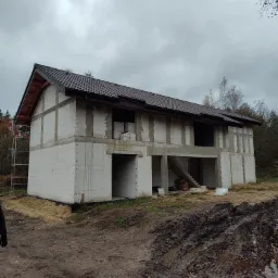 Dwukondygnacyjny dom w stanie surowym otwartym z brązowym dachem, widoczne białe bloczki konstrukcyjne i szare spoiny, teren wokół domu błotnisty z widocznymi śladami opon.