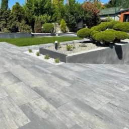 Ułożona szara nawierzchnia z prostokątnych płyt betonowych, z rabatami kwiatowymi i trawnikiem w tle, w słoneczny dzień.