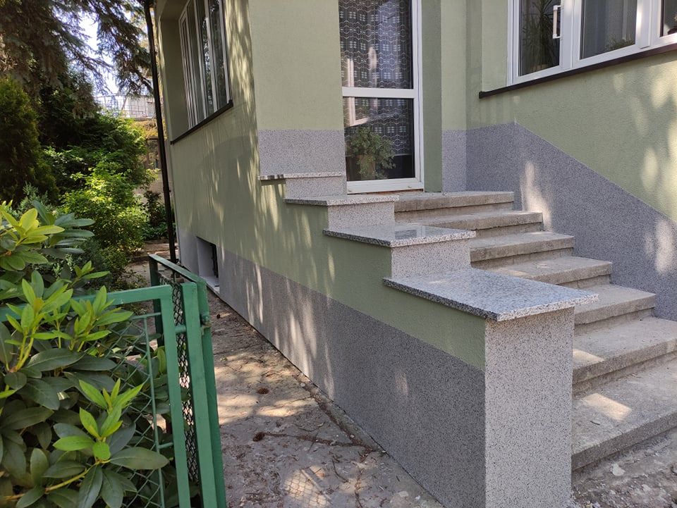 Wykończone schody wejściowe do domu z elewacją w odcieniach zieleni i szarości, z detalami z szarego kamienia dekoracyjnego, widoczne zieleń w tle.
