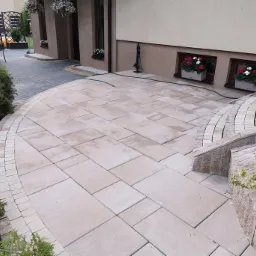 Jasne, prostokątne płyty ułożone na tarasie przed domem, z obrzeżem z małych kostek, widoczne rośliny w donicach w oknach piwnicznych i fragment schod&oacute;w.