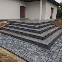 Point-Bud - Nowoczesny taras z szarej kostki betonowej i schodami, prowadzący do domu z jasną elewacją i ciemnym dachem. Widoczne nierówności terenu wokół tarasu.