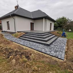 Point-Bud - Nowoczesny dom z tarasem i schodami wyłożonymi szarą kostką brukową, ścieżka z kostki prowadząca do tarasu, pracownik w pomarańczowym kombinezonie układa kolejne elementy, widoczna taczka i paleta...