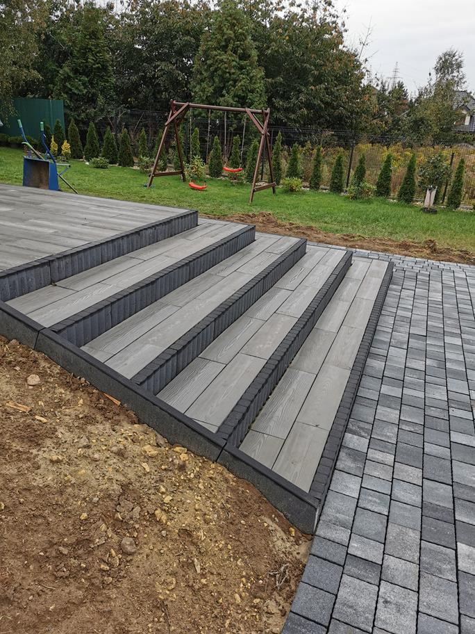 Schody z desek kompozytowych w odcieniach szarości z czarnym obramowaniem, prowadzące z tarasu do ścieżki z kostki brukowej, w tle huśtawka i zielony trawnik.