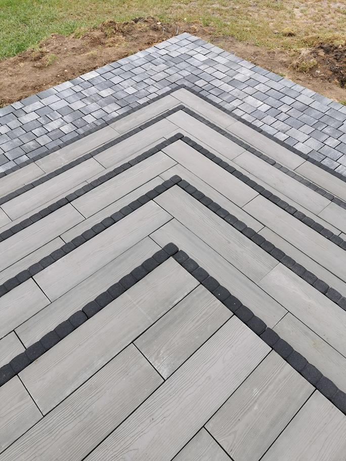 Nawierzchnia z szarej kostki betonowej ułożonej we wzór jodełki, obramowana ciemnoszarą kostką brzegową i przechodząca w kostkę o mniejszym formacie.