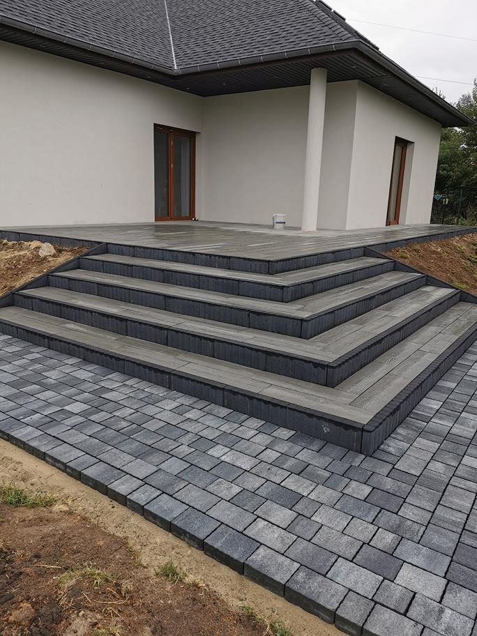 Nowoczesny taras z szarej kostki betonowej i schodami, prowadzący do domu z jasną elewacją i ciemnym dachem. Widoczne nierówności terenu wokół tarasu.