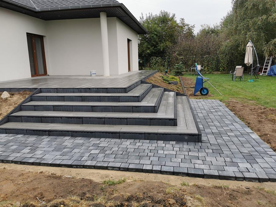 Nowoczesne schody zewnętrzne z ciemnoszarych płyt prowadzące na taras, obok ułożona jasnoszara kostka brukowa, w tle dom z białymi ścianami i ogród z niebieską taczką.