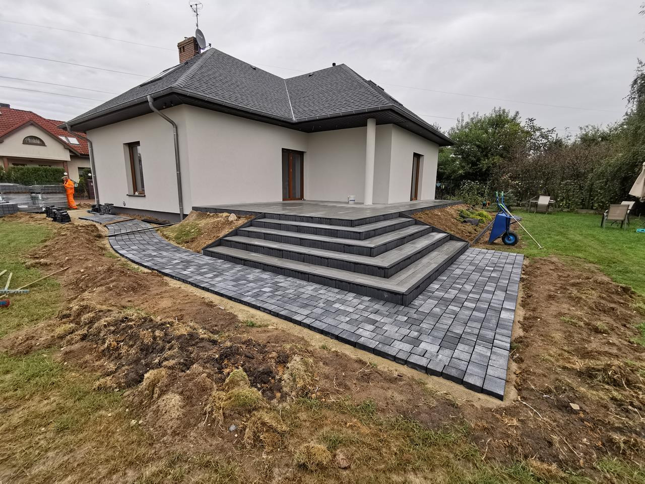 Nowoczesny dom z tarasem i schodami wyłożonymi szarą kostką brukową, ścieżka z kostki prowadząca do tarasu, pracownik w pomarańczowym kombinezonie układa kolejne elementy, widoczna taczka i paleta...