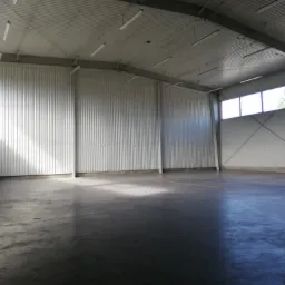 Puste wnętrze magazynu z betonową podłogą, oświetleniem jarzeniowym i ścianami z blachy falistej, z widocznymi oknami w górnej części ściany.