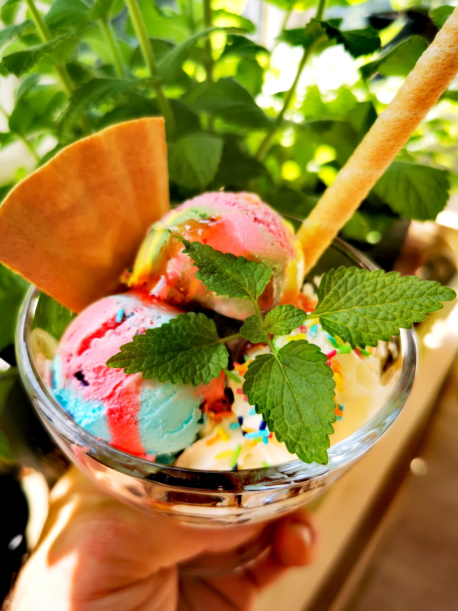 Ręka trzyma deser lodowy w szklanej miseczce z waflem, rurką i listkami mięty na tle zieleni.