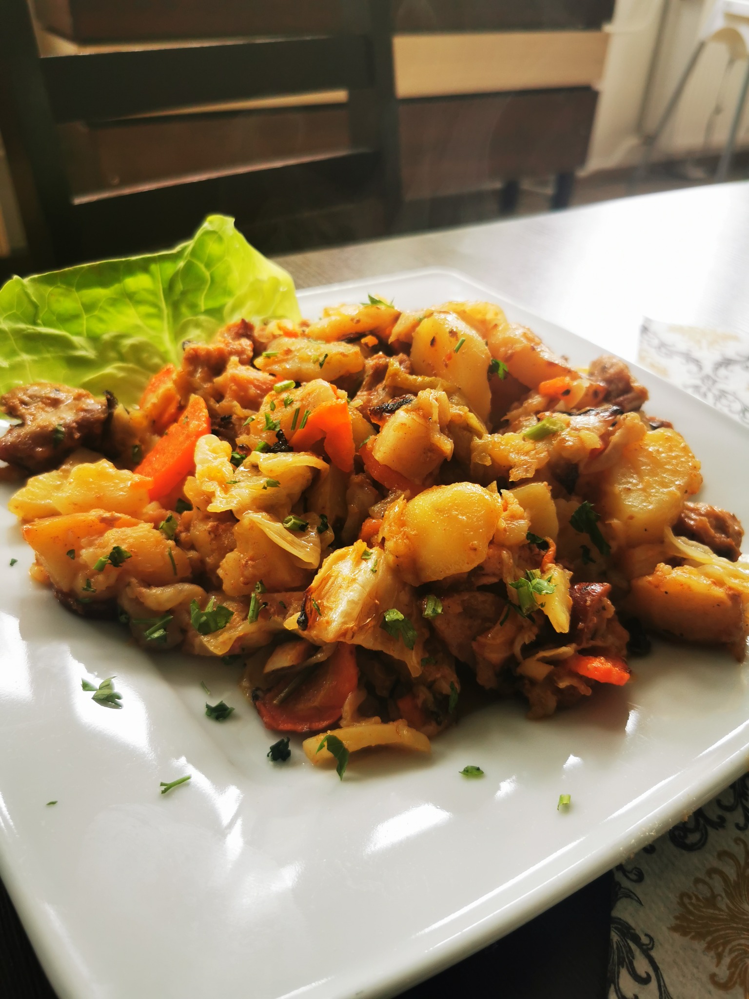 Domowe danie jednogarnkowe z ziemniaków, kapusty, marchwi i mięsa, udekorowane listkiem sałaty i posiekaną zieloną pietruszką, podane na białym kwadratowym talerzu.