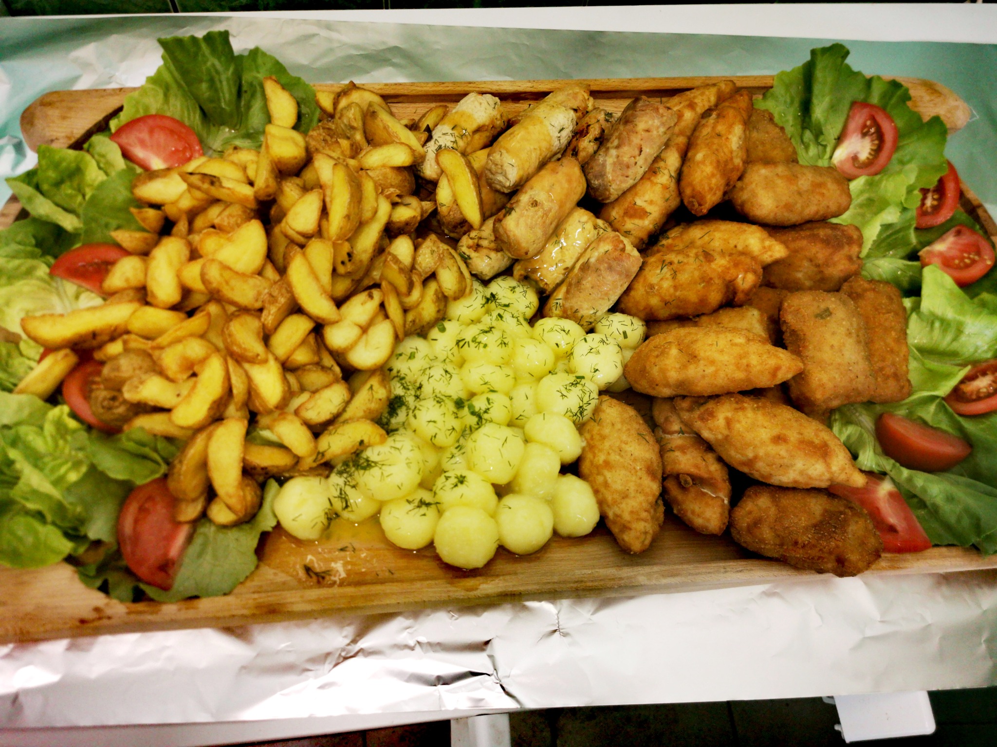 Drewniana taca z różnorodnymi przekąskami: smażone ziemniaki, krokiety, kotlety i gotowane ziemniaki z koperkiem, udekorowana liśćmi sałaty i ćwiartkami pomidorów.