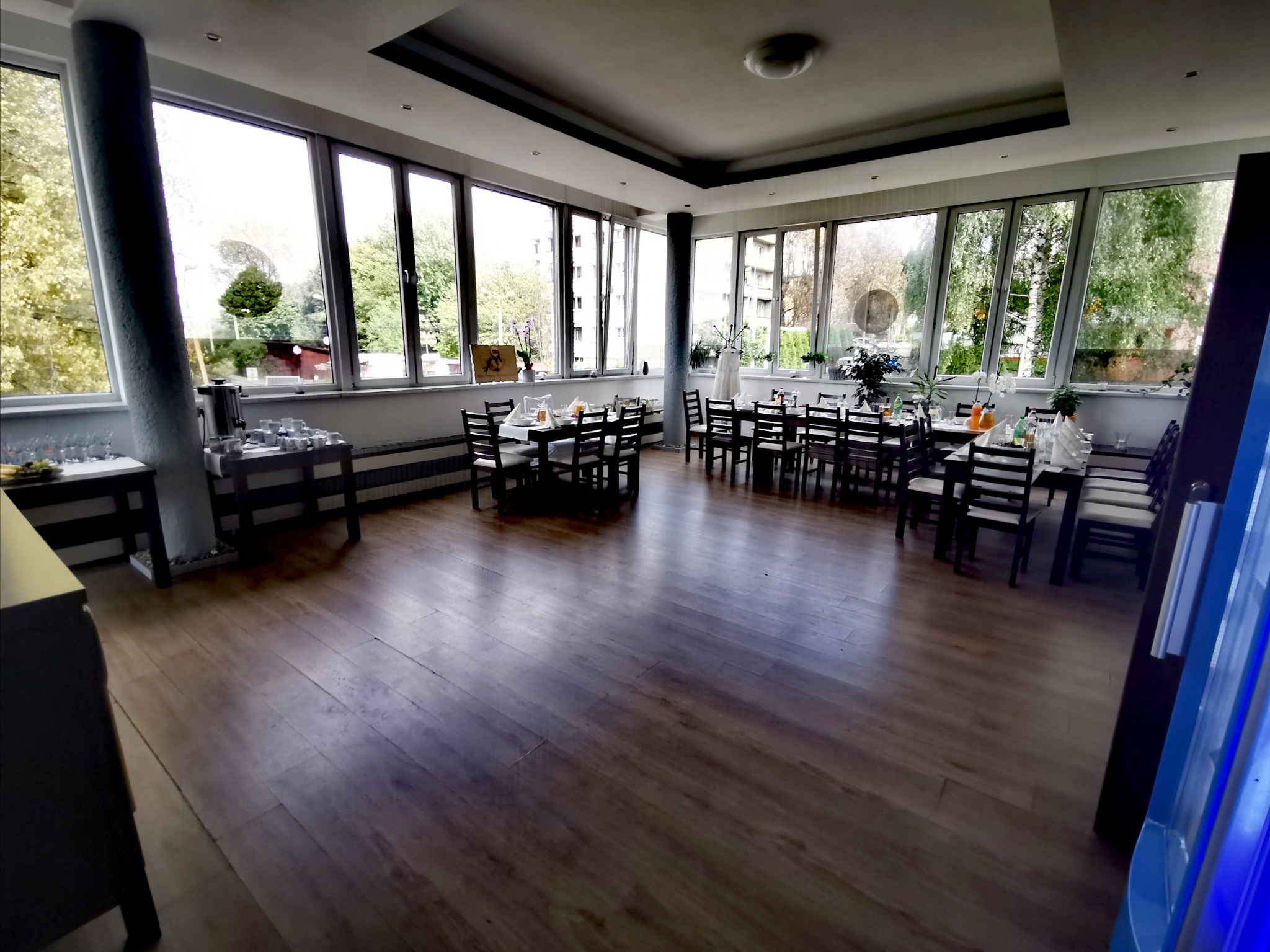 Sala bankietowa z drewnianą podłogą i stołami nakrytymi dla gości, widok zza filaru, duże okna z widokiem na zieleń.