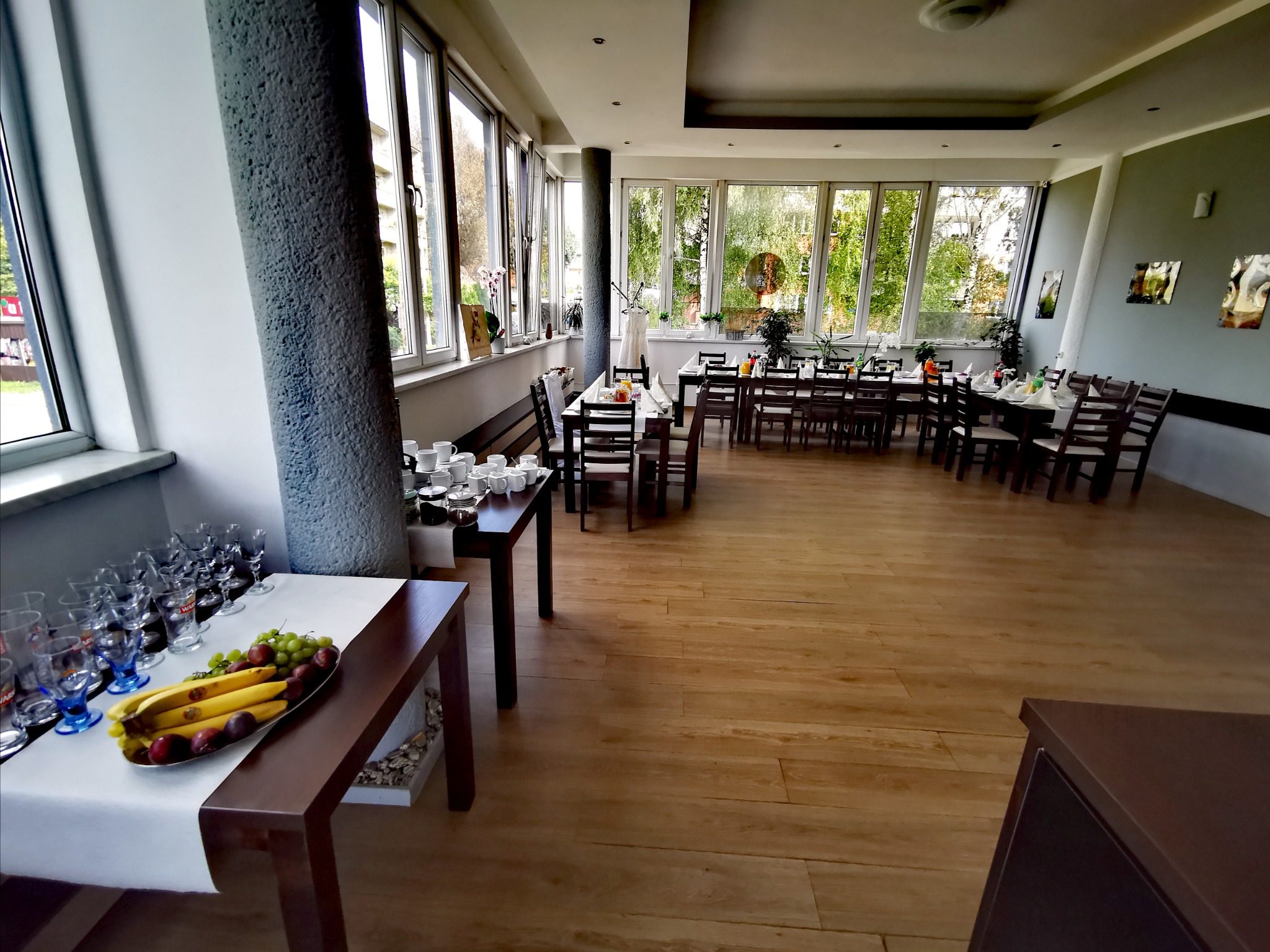 Sala bankietowa z długimi stołami nakrytymi białymi obrusami, przygotowanymi do przyjęcia gości, z widokiem na zieleń za oknami. Na pierwszym planie stół z owocami i szklankami.