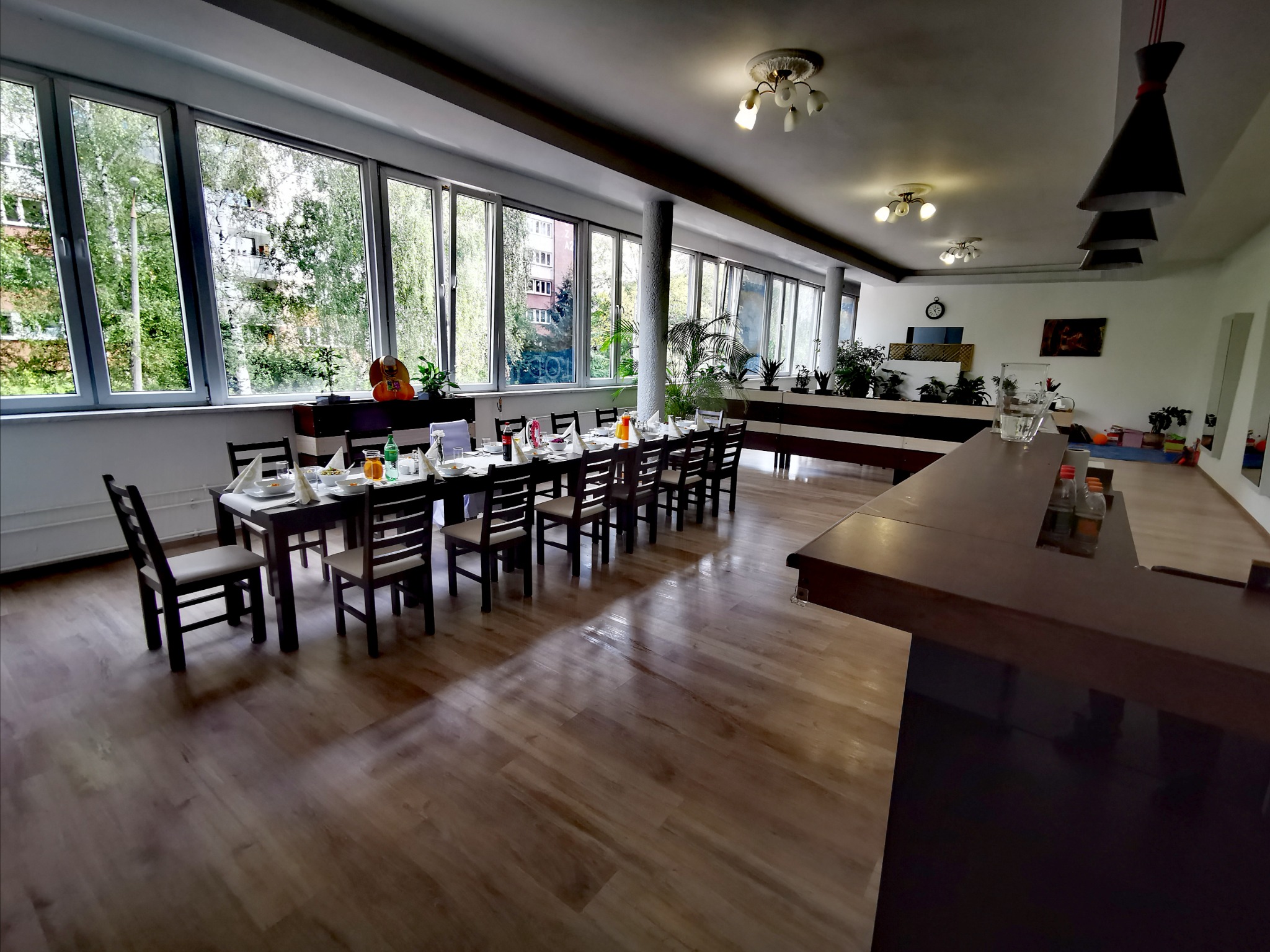 Sala z długim stołem nakrytym do posiłku, widok zza baru, duże okna z widokiem na zieleń na zewnątrz.