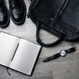 Elegancki zestaw biznesowy: czarne sk&oacute;rzane buty typu brogsy, torba na laptop, notatnik z długopisem i klasyczny zegarek na drewnianym tle.