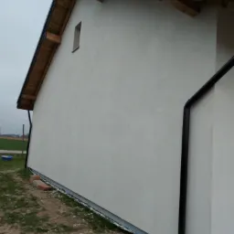 Fragment ściany budynku po nałożeniu jasnego tynku, widoczny fragment dachu z drewnianą konstrukcją, rynna oraz fundament z izolacją przeciwwilgociową, na ziemi resztki materiałów budowlanych i cegły.