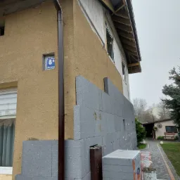 Ocieplanie elewacji budynku jednorodzinnego szarym styropianem grafitowym, widoczna część starej elewacji w kolorze piaskowym, rynna spustowa i numer budynku na ścianie.