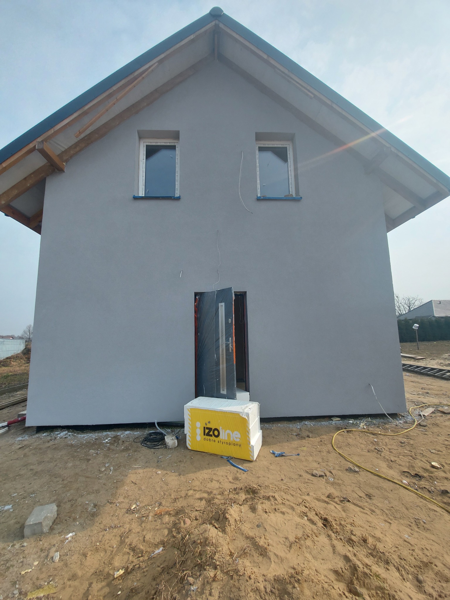 Dom jednorodzinny z szarą elewacją po nałożeniu tynku, widoczne okna z niebieską folią ochronną, otwarte drzwi wejściowe oraz paczka styropianu na ziemi.
